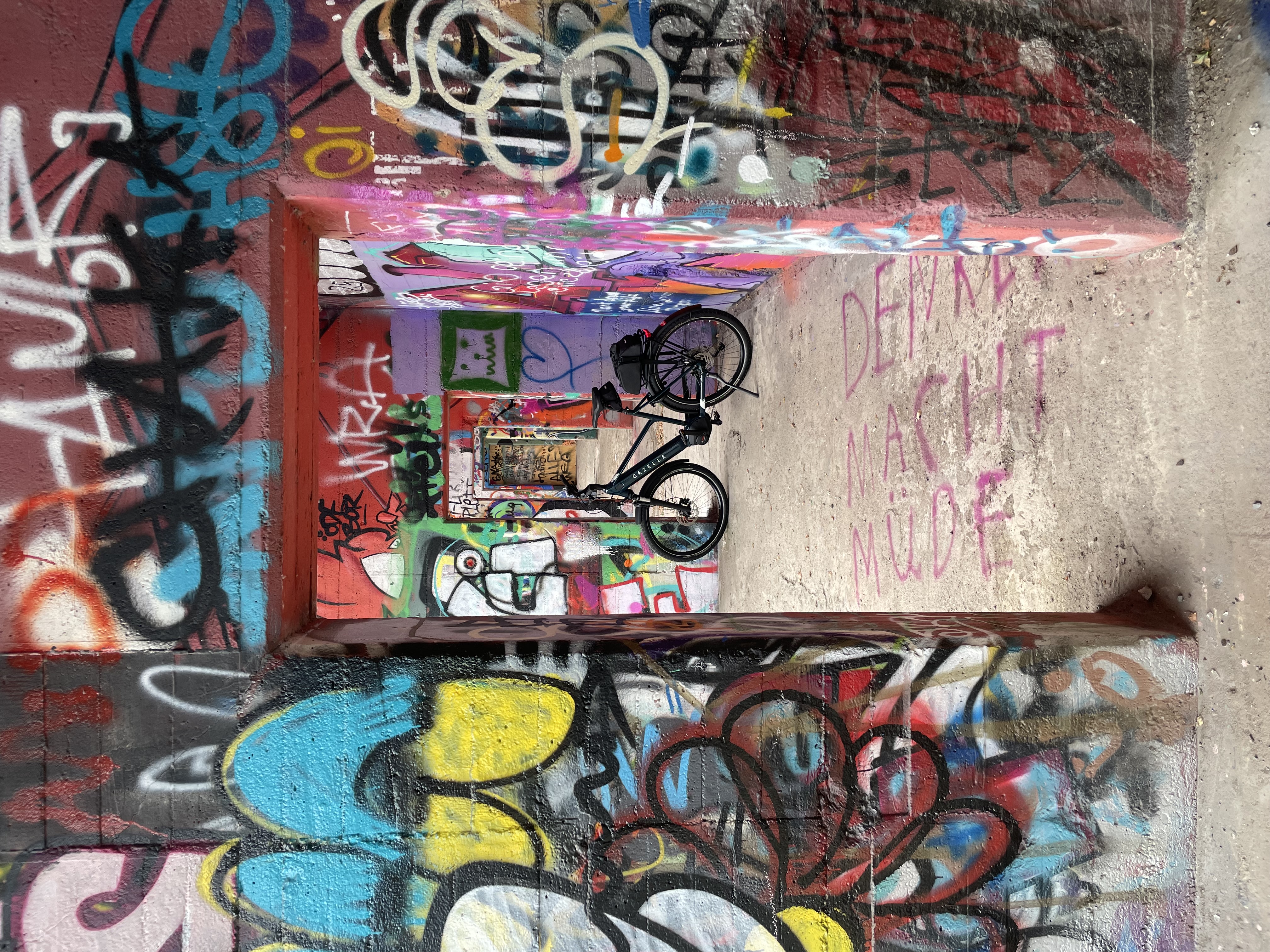 nnenraum einer betonierten, vollständig mit bunten Graffiti bedeckten Ruine; mehrere hintereinander liegende Türöffnungen bilden einen tiefen Blick, in der Mitte steht ein schwarzes Fahrrad.